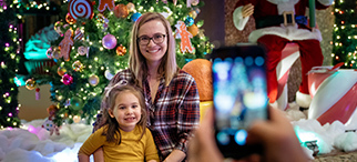 A person taking a picture of a child and a woman with a cell phone on a Christmas setting with Santa Claus and Christmas tree in the background